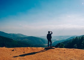 A lone traveler stands on a hilltop, gazing over a vast mountain landscape under a clear blue sky.